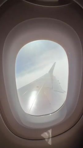 The video shows a view from an airplane window. We see the wing of the plane, with the distinctive pink tail of a Wizz Air aircraft. The wing is partially wet, and sunlight reflects off its surface. Outside the window, there's a view of fluffy white clouds under a blue sky, with a faint outline of land visible on the horizon. The window frame is a light neutral color.