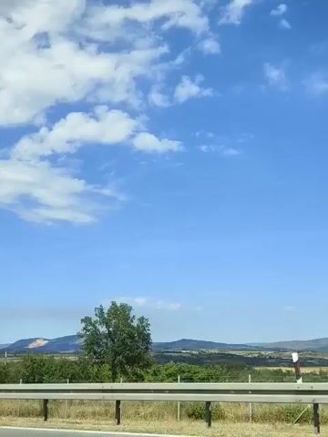 A video shows a blue sky with white clouds and a line of trees and hills in the distance. A metal guardrail is in the foreground, and what appears to be a highway is visible below it. The video is taken from a moving vehicle. There are no people visible in the video.
