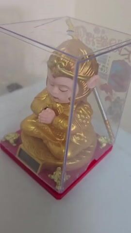 A golden figurine of Sun Wukong, the Monkey King, is displayed in a clear plastic box. The figurine is seated in a meditative pose, with its eyes closed and hands clasped. The box is placed on a white surface, possibly a table or shelf. In the background, a wall is visible. The figurine appears to be a solar-powered toy, as a solar panel is integrated into the base. The overall scene is well-lit, suggesting it was filmed indoors.