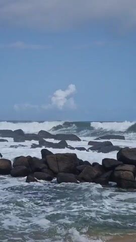 The video shows an ocean scene with waves crashing over dark rocks. The sky is blue with some light, white clouds. The waves are white and foamy as they break against the rocks. The water around the rocks is turbulent and churning. The perspective is from the shore, looking out towards the horizon.