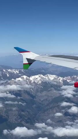 The video captures an aerial view from the window of an airplane. In the foreground, the wing of the aircraft is visible, featuring the Uzbekistan Airways logo on its winglet. Below the wing, a majestic mountain range stretches across the horizon, with snow-capped peaks interspersed with rocky terrain and patches of clouds. The sky is a clear, vibrant blue. The camera appears to be stationary, providing a constant perspective of the breathtaking landscape and the airplane's wing.