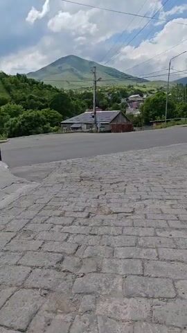 This is a vertical video taken from a low angle looking upwards. The foreground is a cobblestone surface, which appears to be a road or pathway. Beyond the cobblestones is an asphalt road that curves to the right. In the middle ground, there are two houses, one with a white facade and another with a brown wooden exterior. A utility pole stands between them, with wires extending across the frame. Behind the houses, the landscape rises into a lush green hill covered in trees. Further in the background, a large, conical mountain with a light green slope dominates the horizon. The sky is blue with scattered white clouds. The overall impression is a rural setting with a mountain backdrop on a partly cloudy day.