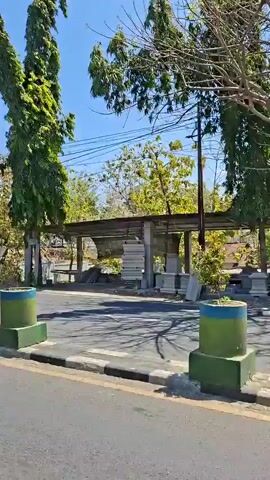 The video begins with a wide shot of a street scene on a sunny day. Tall, slender trees with green foliage line the road, and in the background, there are structures that appear to be part of a covered waiting area or bus stop. A blue and white bus is parked on the left side of the road, near a green and blue concrete barrier. A road sign is visible on the right side of the frame.

The camera pans and shifts focus, showing different parts of the street. A bright yellow car is seen driving away, and its side is partially visible. Another shot reveals a bare-branched tree in the middle of the scene, with a small blue structure behind it.

Further along the road, another yellow car is parked. The camera then follows a motorcycle with two people on it, moving away from the camera. The person in front is wearing a helmet and a dark jacket, and the person behind is also wearing dark clothing. They are riding on the road, passing by a low white wall with green decorative elements. The video ends with a shot of the motorcycle moving further down the street, with trees and power lines visible in the background. The overall environment appears to be an urban or suburban area with moderate vegetation.