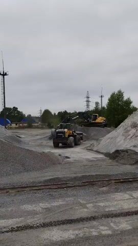 The video shows a construction site or quarry under an overcast sky. The foreground has piles of gravel and a set of railroad tracks. In the middle ground, a yellow Volvo loader is stationary, facing forward. An excavator with a black arm is actively digging into a pile of gravel and loading the loader. Further in the background, there's another yellow excavator parked on a higher pile of gravel. Trees, utility poles, and a building with a blue roof are also visible in the background.