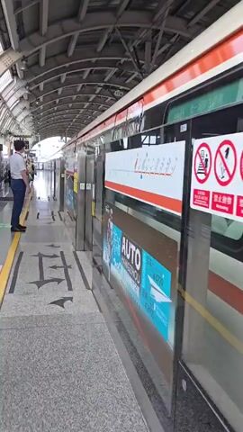 A man wearing a white shirt and blue pants stands on a train platform, facing away from the camera and towards the train. The train is on the right side of the frame, with its doors closed. The platform is covered by a metal roof with visible support beams and lighting fixtures. The floor of the platform is paved with speckled gray tiles, marked with yellow and green lines, and arrows indicating directions. There are several large advertisements on the side of the train. In the background, other people are visible on the platform, and the train station appears to be busy.