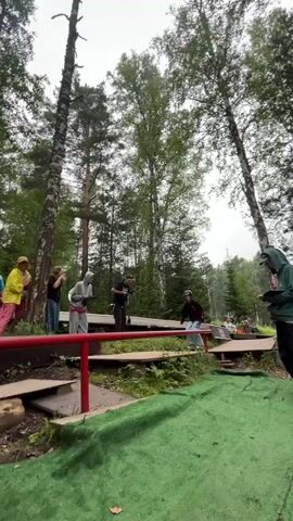 The video shows a group of people gathered in a forest setting, possibly at an outdoor park or event. In the foreground, a person on roller skates is attempting to balance on a narrow, elevated beam painted red. They are wearing a helmet and light-colored pants. The beam appears to be part of an obstacle course or a recreational area.

Behind the person on skates, several individuals are watching. Some are standing on a wooden platform next to the beam. One person is wearing a yellow raincoat, another a blue shirt, and others are in casual clothing. The environment is lush with tall trees and green foliage, suggesting a wooded or park area. The ground is covered with grass and some wooden logs.

As the video progresses, the person on skates attempts to navigate the beam, demonstrating their balance and coordination. The camera follows their movement, with a brief, blurry shot of someone holding a dark object in the foreground at one point. The overall atmosphere seems relaxed and recreational, with spectators observing the skating activity.