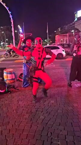 Three costumed performers dance in a city square at night. The primary focus is a Deadpool character in the center, flanked by Spider-Man variants. One Spider-Man, in a cowboy hat and blue harem pants, stands to the left, and a black-suited Spider-Man stands to the right. They are dancing on a red brick surface near a street, with cars and city lights in the background. The lighting is predominantly pink, highlighting the dancers' bright costumes. A speaker and some bags are located to the left, indicating the source of the music for their performance. The Obelisco de Buenos Aires is faintly visible in the distance.
