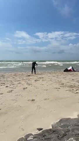 A person in a black wetsuit stands on a sandy beach, holding a white rope that extends into the choppy ocean. A colorful kite, black with red and white accents, lies on the sand near the water's edge. The sky is blue with scattered clouds.