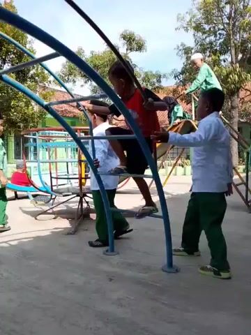 The video shows a school playground with children playing. Several boys dressed in white shirts and green pants are on and around a blue metal climbing structure. One boy in a red shirt is climbing on top. Another boy is standing nearby, possibly assisting or watching. There are other children on the ground, some in similar attire and some in green dresses and hijabs. The background shows a building with green and white pillars, windows, and a terracotta-tiled roof. Some of the girls are sitting on the floor of the covered walkway in front of the building. The overall setting is a sunny day at a school.