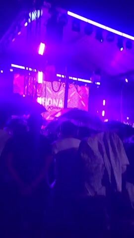 A group of people are gathered at an outdoor event under a tent, illuminated by vibrant pink and blue lights. It appears to be raining, as many attendees are holding umbrellas, some of which are dark and some lighter colored. A stage with lighting equipment is visible in the background, suggesting a performance is taking place. Some individuals are holding phones up, likely recording the event. The overall atmosphere seems lively despite the rain.