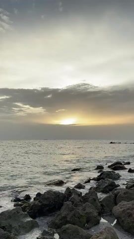 A rocky shoreline meets the ocean under a cloudy sky at sunset. The sun is a bright glow partially obscured by clouds, casting a warm light on the water and sky. Waves gently break against the dark, jagged rocks in the foreground. The water is choppy and reflects the muted colors of the sunset. The overall atmosphere is serene and tranquil.