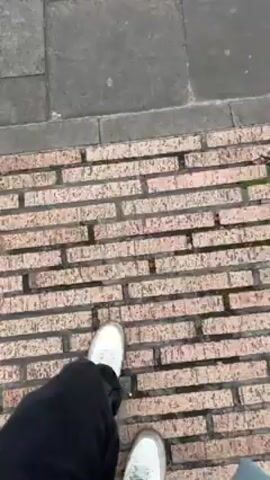 The video shows a person walking on a sidewalk in a city, stepping from a brick walkway to a sidewalk made of gray stone tiles. The camera follows their feet as they walk. As the person turns and walks forward, another person in a brown jacket walks past them, carrying an umbrella. The camera pans up to show the surrounding city, including a modern plaza and tall buildings. The camera then continues to pan up to show stairs leading up to a stone building and other people on the sidewalk. The sky is cloudy.