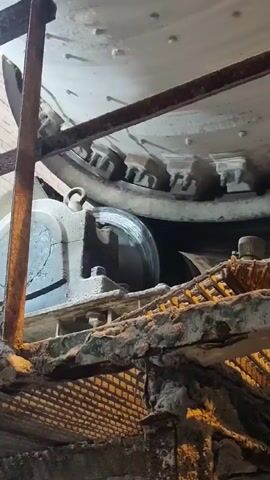 The video shows a close-up view of industrial machinery. The primary object is a large, light-colored, cylindrical structure with a toothed rim, possibly a part of a mill or grinding machine. It is supported by a rusty metal framework. Below this large cylinder, a smaller, dark-colored cylindrical component with metal banding is visible, which appears to be a roller or wheel. This roller is mounted on a rusted metal platform, which is further supported by a grated metal walkway. The overall setting suggests an industrial environment, possibly a factory or processing plant, with visible wear and tear on the metal structures, indicating age and use. Dust and debris are present on the surfaces. The lighting is artificial and somewhat dim, casting shadows and highlighting the textures of the machinery and the environment. There is no visible text in the video. The focus is on the mechanical components and their weathered state. The activity appears to be the static observation of this machinery, as no movement is depicted beyond what might be subtle vibrations of the structure itself.