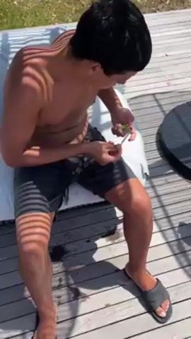 A shirtless person sitting on a cushion on a wooden deck lights a rolled cigarette or joint with a green lighter. The person wears dark-colored shorts and gray sandals. A small round table sits to the side. The scene is outdoors in bright daylight, with shadows cast by a slatted awning.