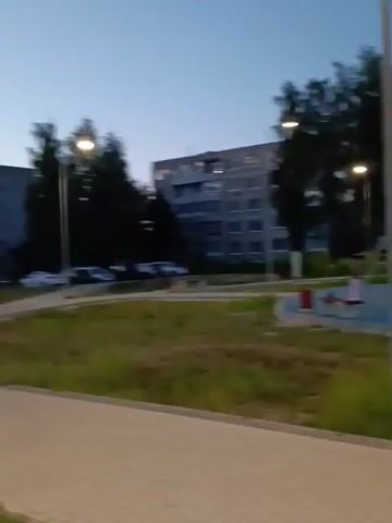 This is a handheld video clip of a playground area outside of an apartment building in Sudogda, Russia. The sky is a clear, deep blue, indicating it is late evening or early morning. The sun has not yet risen or has already set, as artificial lights are visible on the playground and on the building, illuminating the scene.

The playground features several pieces of equipment, including a colorful climbing structure with blue, yellow, and orange elements, a red and white swing set, and a red trash can. There appear to be at least two individuals on the playground, one on the climbing structure and another near the swings. They are too far away and the image quality is too low to discern any specific details about them or their expressions.

In the background, a multi-story apartment building dominates the landscape. Its facade is grey with numerous windows. Cars are parked in front of the building and on the street. A paved walkway runs through the foreground of the video, bordered by patches of green grass and some weeds. The overall atmosphere is quiet and peaceful. There is no visible text in the video.