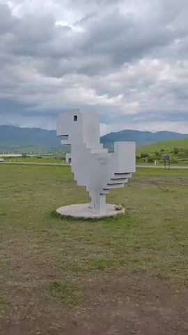 The video shows a 3D pixelated statue of a dinosaur in a grassy field. The statue is white and made of concrete or similar material. It resembles the dinosaur from the Google Chrome offline game. The sky is overcast with dark clouds. In the background, there are rolling hills and a distant mountain range. The grass is green but patchy in places. There is a small road and some trees visible in the background. The camera pans slightly around the statue.
