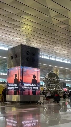 The video shows the interior of a modern airport terminal. In the foreground, a large, cubical structure stands, adorned with advertisements for Soekarno-Hatta International Airport. The advertisements feature silhouettes of people against a sunset backdrop and the airport's logo. To the right, a large, shiny, balloon-like sculpture of a cartoon character is visible. Several people are walking through the terminal, some with luggage, suggesting travel activity. The floor is highly reflective, mirroring the ceiling and the objects within the space. The ceiling itself is a distinctive, geometric design with numerous light panels. The overall atmosphere is bright and spacious.