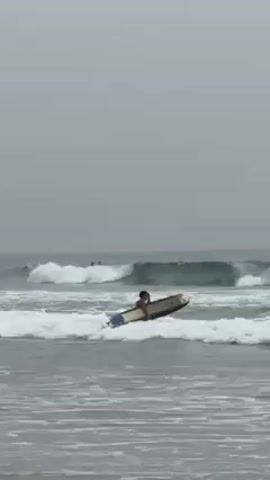 The video shows a surfer kneeling on a surfboard in the ocean, with waves breaking behind him. There are other surfers visible further out in the water. The sky is overcast. A child walks along the beach in the foreground. The surfer transitions to a sitting position on the board.