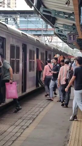 A train is stopped at a station platform, with doors open. Several people are visible, some boarding or disembarking the train, while others are waiting on the platform. The train is light-colored with purple accents, and has several windows and doors. The platform is made of concrete and has yellow markings. In the background, there is a covered section of the platform with signage and a building. A few people are carrying bags or luggage. The overall scene depicts a busy train station environment.