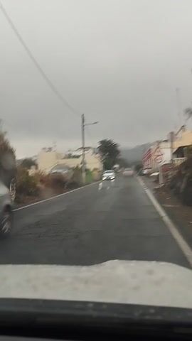 The video shows a POV shot from inside a vehicle driving on a wet road. The road appears to be in a town or village, with buildings lining both sides. The sky is overcast and the weather appears to be rainy, contributing to the wet road conditions. The time of day is difficult to determine due to the overcast sky, but it seems to be daytime.

Several cars are visible on the road, some parked and some driving. In the distance, a white car is driving ahead, its headlights illuminated. A pedestrian crossing sign with a speed limit of 40 is visible on the right side of the road. The buildings are of various colors and styles, some appearing to be residential homes and others possibly businesses. Palm trees are also visible, suggesting a warm climate.

The overall mood of the video is somewhat muted due to the weather and the gray sky. The wet road and the movement of the car create a sense of travel. There are no people clearly visible, though the pedestrian crossing sign implies their presence in the area. The scene is set in a relatively ordinary street environment, with elements of rural or semi-urban life. The location context of Arucas, Spain, suggests a possible Mediterranean or Canary Islands setting. There is no discernible text other than the number "40" on the speed limit sign.