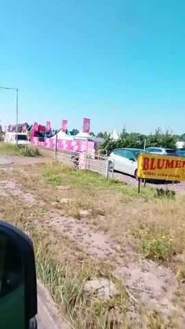 The video shows an outdoor event with multiple tents and inflatable structures set up. The tents are white and yellow with pink banners and flags. The inflatable structures are colorful, with one appearing to be a large archway. Several cars are parked in the background, and there is a fence running along the perimeter of the event area. The ground is covered in grass and dirt, and the sky is clear and blue. The camera appears to be moving, possibly from a vehicle, as the scene is slightly shaky.