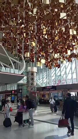 The video captures a busy airport terminal with a large art installation of golden squares hanging from the ceiling. Several travelers are visible, each with luggage. Some are walking towards the camera, while others are moving away. The overall atmosphere is one of transit and movement within a modern, well-lit public space. The installation creates a dynamic visual effect, reflecting light and adding a touch of glamour to the terminal's architecture.