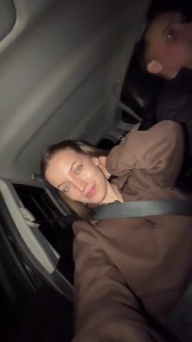 A young woman sits in the passenger seat of a car at night, wearing a brown sweatshirt and a seatbelt. She is blonde with blue eyes and a neutral expression. A man is in the driver's seat; his face is visible in the dim light. The car interior is dark. A baby car seat is visible in the back of the car. Music plays in the background.