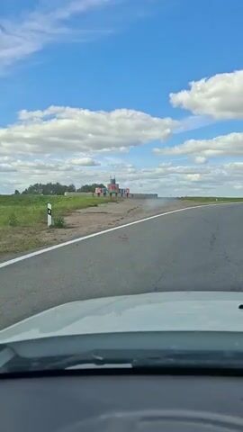 The video captures a view from a vehicle moving along an asphalt road. The road has a white line marking its edge. To the left of the road, there is a grassy field with some bushes or trees in the distance. In the middle ground, a structure resembling a castle or fortress is visible. It appears to be made of colorful blocks, possibly shipping containers, arranged in a multi-layered formation with towers and flags. In front of this structure, there are several large, cylindrical objects, which might be hay bales or large tanks. The sky is blue with scattered white clouds, suggesting a daytime setting with fair weather. The camera is positioned inside the vehicle, showing a portion of the dashboard and windshield in the foreground. The car is moving forward, and the castle-like structure is receding into the distance.