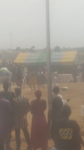 The video displays an outdoor scene captured during what appears to be a lively gathering or event. The ground is sandy or dusty, and a noticeable haze permeates the air, suggesting either dust or atmospheric conditions, under bright daylight.

Numerous people are visible throughout the frame, primarily in the mid-ground and background. They are standing, walking, and interacting, creating a bustling atmosphere. In the background, a large, tent-like structure with yellow and green stripes is prominent, serving as a focal point for the event.

In the foreground, a tall, slender pole stands vertically on the left side. To the right, several individuals are closer to the camera. One person is wearing a striking red garment. Another person on the far right has a dark piece of clothing or an item with the word "CAUTION" clearly visible in yellow lettering. In the mid-ground, near the tent, a light-colored object, possibly a ball, is momentarily suspended in the air, indicating some activity or game might be taking place. Other people are dressed in various attire, including striped shirts, and some appear to have bags or packages on the ground near them. The overall scene conveys a sense of an active and well-attended public event.