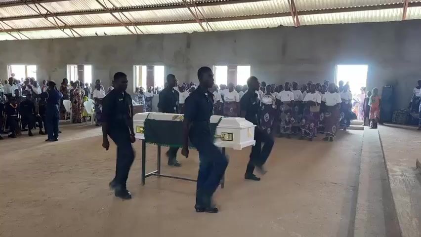 The video shows a funeral service taking place inside a large, open building with a corrugated metal roof and concrete walls. The floor is dirt. In the foreground, four individuals dressed in dark blue uniforms stand at attention. In front of them, a white casket adorned with a green and gold cloth rests on a metal stand. A crowd of people, some in white and others in colorful traditional attire, are gathered in the background, facing the casket. Several individuals in uniform are seen near the casket, appearing to prepare for its removal. To the left, a group of people in dark uniforms are seated, some with instruments resembling drums. To the right, more attendees are standing. Sunlight streams in from open doorways at the far end of the building, illuminating the scene. The overall atmosphere suggests a solemn occasion.