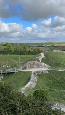 A scenic video shows a panoramic view of an ancient landscape, likely castle ruins or a hillfort, under a partly cloudy blue sky. The camera slowly pans from left to right, revealing rolling green grassy mounds and fields in the distance. In the foreground, there are visible stone remnants of old walls and structures, suggesting historical significance. A wooden bridge with railings spans a ditch or low area between two grassy sections. Lush green trees and bushes are scattered throughout the scene, particularly on the edges of the mounds and in the background. The overall impression is one of a vast, historical, and well-preserved outdoor site, with a mix of natural beauty and man-made structures. No people or visible text are present in the video.