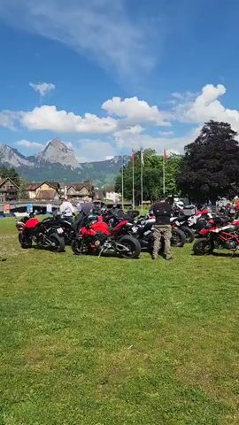 The video shows a group of motorcycles parked on a grassy field. People are standing around the motorcycles. In the background, there's a lake surrounded by mountains. The sky is blue with some clouds. Some people are walking along the lakeshore. There are trees and flags visible. The scene appears to be a motorcycle gathering or event in a scenic location.