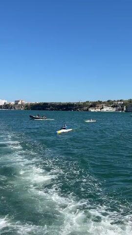 The video shows a wide expanse of blue water with several people on paddleboards and kayaks spread across it. The water is rippled and shows a foamy wake at the bottom left of the screen, indicating movement. In the background, a rocky coastline is visible, lined with buildings and trees. A few boats are also present on the water, and some larger yachts are moored in the distance. The sky is a clear, bright blue. The scene appears to be a recreational area, possibly a bay or harbor, on a sunny day.