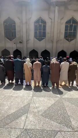 A group of men are praying outside a building. The men are standing in rows, facing away from the camera. They are all wearing traditional clothing. The building appears to be a mosque. The ground is paved with tiles. The lighting suggests it is daytime.