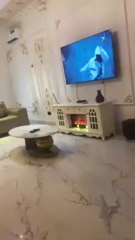 The video depicts a living room with white marble flooring and ornate white and gold wall decorations. A beige couch sits on the left side of the frame, facing a circular coffee table with a marble top and gold base. A television is mounted on the wall, showing a person wearing white. Below the TV is a white media console with a simulated fireplace effect. The video pans across the room, providing a view of the furniture and decor. Music plays in the background.