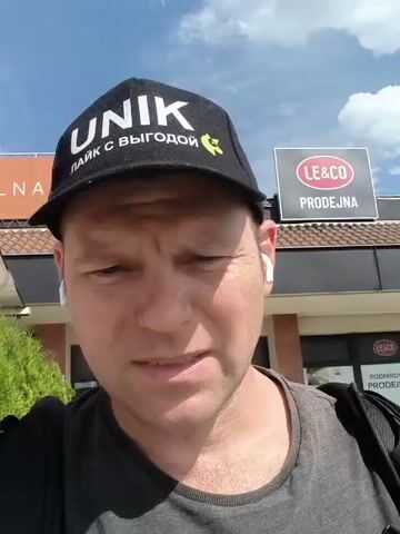 A man is featured in a close-up, chest-up shot, appearing to record a video outdoors on a sunny day. He is wearing a dark t-shirt, a black baseball cap with "UNIK" and "ЛАЙК С ВЫГОДОЙ" (LIKE WITH PROFIT/ADVANTAGE) written on it in white and yellow, and white earbuds. He has short, fair hair and a slightly serious expression. In the background, under a bright blue sky with scattered clouds, is a commercial building. Prominently visible signs on the building include "LE&CO PRODEJNA" (PRODEJNA means "shop" or "store" in Czech) and "PODNIKOVÁ PRODEJNA" (Company Shop/Factory Shop). Another sign is partially visible with "NA". The man is speaking, likely addressing the camera, with a slight furrow in his brow. He is wearing a dark backpack, the strap of which is visible on his right shoulder.