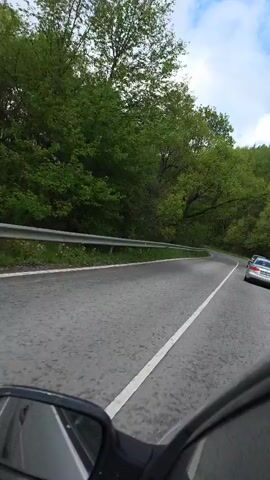 The video shows a winding road through a forest. The road is asphalt and has a white line dividing the lanes. There is a guardrail on the left side of the road. The forest is dense and green. Two cars are visible driving on the road. The camera appears to be mounted on a motorcycle, as part of the motorcycle's side mirror is visible in the lower left corner of the frame. The sky is partially visible with some clouds.