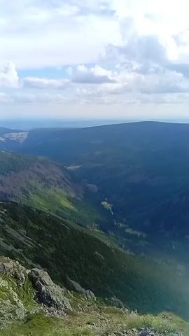 The video shows a panoramic view of a mountainous landscape on a cloudy day. The camera is positioned high up, looking down into a deep valley. The valley is covered in dark green trees, with some areas appearing sparser or showing signs of deforestation. A thin, winding river or stream can be seen in the distance within the valley. The slopes leading down into the valley are steep and rocky, with some patches of green grass and scrub. In the foreground, the ground is covered in rocks and short green grass. The sky above is partly cloudy, with patches of blue sky visible. The overall impression is a vast, wild, and rugged natural environment. There are no people or man-made objects visible in the video.