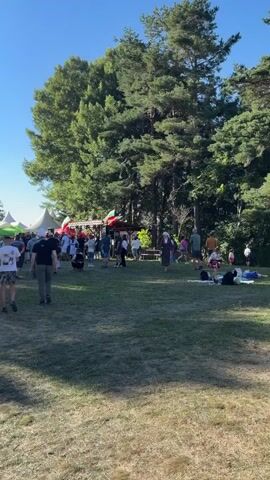 The video shows a wide shot of an outdoor event, possibly a festival or market, on a sunny day. A clear blue sky is visible above tall, green trees. In the foreground, a grassy field is dotted with shadows from the trees. Several people are walking across the field, some in casual clothing. In the mid-ground, there are numerous tents, some white and some green, set up in rows. A stage or performance area is visible behind some of the tents, with people gathered in front of it. Various flags, appearing to be red, white, and green, are displayed. More people are milling around the tents and near the stage. To the right, a few people are relaxing on the grass. The overall atmosphere appears to be relaxed and festive.