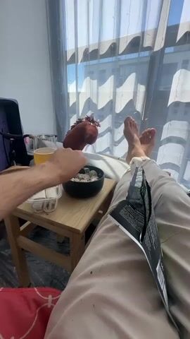 A person is reclining on a bed with their feet up, holding a fork in their right hand. In front of them, on a wooden side table, is a bowl of dumplings, a glass of amber liquid, and a plastic container with what appear to be small wrapped food items. To the left of the table, a purple suitcase or bag is partially visible. A toy or puppet, resembling a bird with a brown head and red accents, sits on the bed. The person's left hand is reaching towards the bowl of dumplings with a spoon. The room has large windows with sheer curtains, and daylight is streaming in, casting shadows from the window blinds. The person is wearing light-colored pants, and a black and white magazine or booklet is resting on their lap. The scene suggests a casual moment of relaxation and eating.