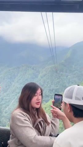 The video shows several people inside a cable car with a scenic mountainous view. Two young women in the foreground are looking at their phones, one capturing a photo or video of the landscape. A young man in a white cap sits next to them, also looking at his phone. Another young man in a white t-shirt is visible further inside the cable car, looking out the window. The cable car appears to be moving over a lush, green, and mountainous terrain. The sky is overcast, adding a dramatic effect to the scenery.