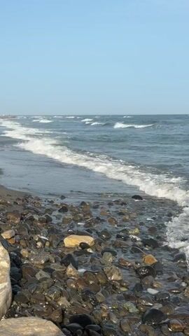 The video shows a stony beach with waves rolling in from the sea. The water is a deep blue, and the sky is a clear, light blue. There are no people visible in the video. The camera is positioned low, close to the ground, giving a close-up view of the pebbles and the water. The waves are gentle, with white foam crests. The overall impression is one of peacefulness and natural beauty.