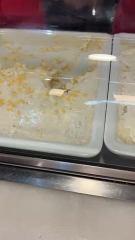 The video pans across a refrigerated display case filled with various food items. On the left, there are two white rectangular trays containing what appears to be creamy salads or dips. The first tray is topped with yellow flecks, possibly corn or cheese. The second tray contains a similar creamy mixture with visible pieces of red and white ingredients, and two shrimp. To the right, a metal compartment holds two white root vegetables, possibly turnips, and a large, textured, brown object. Further to the right, another white rectangular tray holds a salad of chopped white ingredients, likely onions or potatoes, mixed with herbs and a light dressing. A person wearing a red shirt is partially visible in the background, and the display case has a glass front. The overall environment suggests a food service establishment, possibly a deli or buffet.