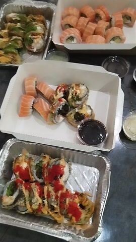 The video displays an overhead view of multiple containers filled with various types of sushi, suggesting a take-out or delivery meal. The scene is set indoors, likely on a dark table or counter.

In the upper left, a white cardboard box holds numerous sushi rolls predominantly wrapped in thin slices of salmon, with a light-colored filling, possibly cream cheese and avocado. Below this, an aluminum foil tray contains deep-fried sushi rolls, some topped with green herbs or sauces, and appearing golden-brown.

In the center, another white cardboard box showcases a mix of sushi: some appear deep-fried with various toppings, while others are salmon-wrapped rolls similar to those in the first box. This box also includes a small plastic cup filled with dark soy sauce.

To the right, partially visible in an aluminum foil tray, are more sushi rolls, many adorned with bright red toppings or sauces, giving them a vibrant appearance.

Several small, clear plastic cups, likely containing condiments such as ginger or wasabi, are scattered among the containers. The camera gently pans across the different food items, offering a closer look at the variety of sushi prepared for consumption. There is no visible text on any of the packaging.