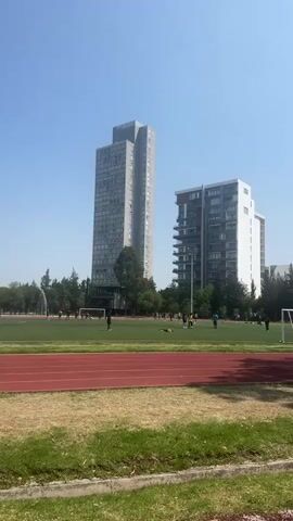 The video shows an outdoor soccer field with a red running track around it. There are multiple people on the soccer field, some of whom appear to be kicking a ball. In the background, there are two tall buildings, one grey and rectangular, the other a more modern white building with balconies. The sky is blue and clear. The foreground consists of a grassy area with some dead grass.