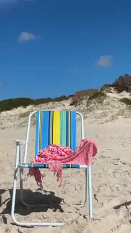 The scene captures a vibrant beach chair set against a backdrop of sand dunes under a clear, blue sky. The chair is positioned on the sand, with a pink patterned beach towel draped over its striped seat. The stripes on the chair are colored blue, yellow, and green. The white-framed chair faces the camera, and behind it, sand dunes rise, dotted with scrubby green vegetation. The sky above is a bright azure, sparsely populated with a few small, fluffy clouds. The lighting suggests a sunny day.