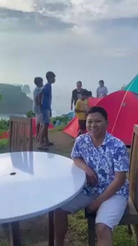 The video shows a man sitting at a white table and smiling at the camera. Behind him, several people are walking around a campsite with red and green tents set up on a grassy hill overlooking a beach. The scene is set against a backdrop of a cloudy sky and ocean with rocky outcroppings. As the camera pans, a young girl walks into the frame.
