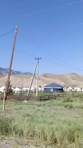The video shows a view of a village consisting of yurts, set against a backdrop of rolling hills and a clear blue sky. In the foreground, there is tall grass and a wooden fence. A wooden utility pole with wires is prominent on the right side of the frame. The camera pans to the right, revealing an arched entrance to the village, adorned with a circular emblem. More yurts are visible behind the fence and entrance, and the landscape continues with sandy hills and distant mountains. The overall impression is of a rural, possibly nomadic, settlement in a dry, mountainous region.