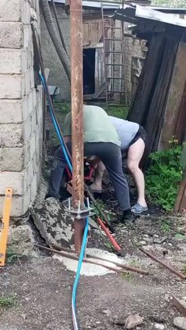 The video shows two men working together to install a metal pole near a corner of a brick building. The man on the left is wearing black pants and shoes, and the man on the right is wearing dark shorts and sandals. Both men are bending over, working on the pole, which is held in place by a metal bracket and several bolts. A blue and white wire runs down the pole. A level is resting on the ground, and a ladder is visible in the background leaning against a shed.  The ground is covered with dirt, rocks, and debris. The men are using a tool that is making a buzzing sound.