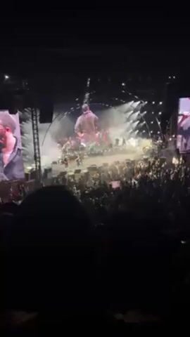 The video shows a performance on a large stage with an audience. The stage features a band and several performers, with large screens showing close-ups of the main singer. There are curved light structures around the stage. The audience is densely packed, and many people are standing. Towards the front, a group of people are holding up a pink sign with text and an image, possibly a memorial or tribute. The lighting is bright and focused on the stage.